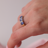 Close-up of a hand wearing a ring with blue sapphires and diamonds on a neutral background.