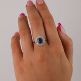 18ct white gold sapphire halo dress ring worn on hand showing rich blue gemstone and sparkling diamond surround.